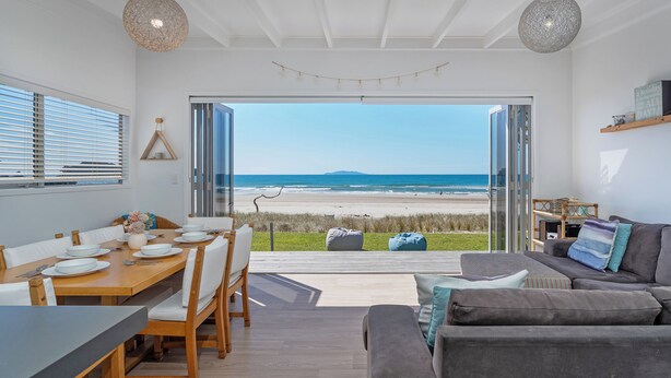 A modern living room with a large glass door leading to a beach view, a dining area with a wooden table and chairs, and a grey sofa with cushions. - Beachfront Beauty - Whangamata Beachfront Home (Whangamata)