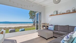 A modern living room with a large glass door leading to a balcony overlooking a beach. - Beachfront Beauty - Whangamata Beachfront Home (Whangamata)