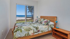 A bedroom with a wooden bed, a nightstand, a blue lamp, and a view of the beach through large windows. - Beachfront Beauty - Whangamata Beachfront Home (Whangamata)