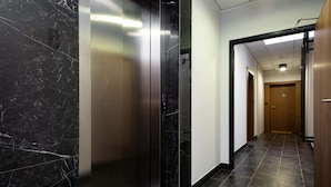 A modern hallway with a glass elevator, dark marble walls, and a tiled floor.