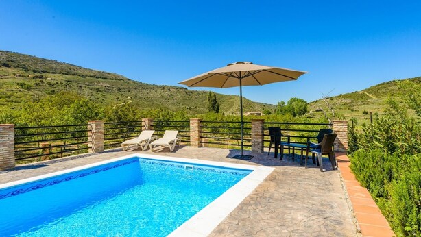 A swimming pool with lounge chairs and an umbrella, surrounded by a stone deck and a metal fence, with hills in the background. - Los Linarejos Ronda by Ruralidays (Málaga)