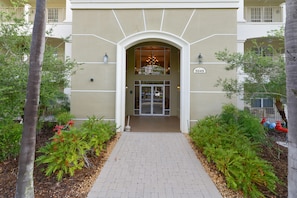 A building entrance with a brick pathway, surrounded by greenery and palm trees.