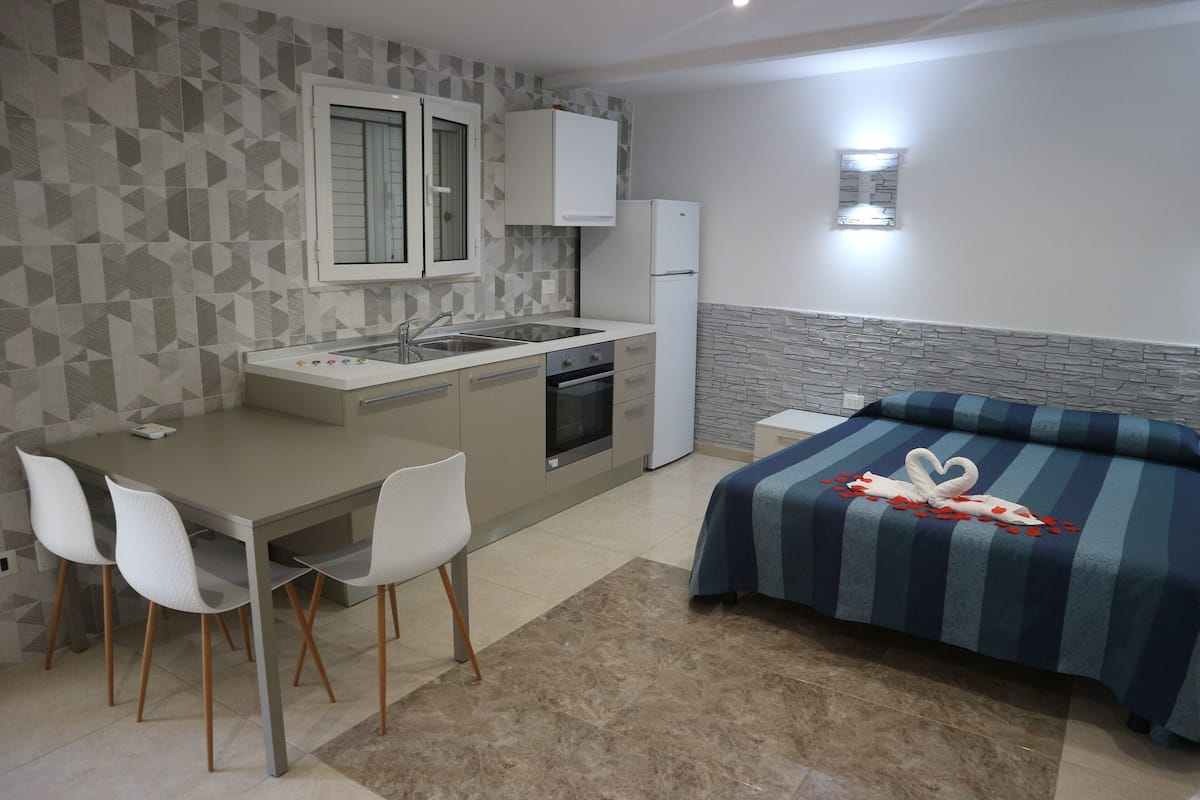 A compact hotel room with a kitchenette, a bed with a blue striped bedspread, and a small bedside table.
