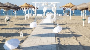 Decoração de casamento na praia, com um caminho que leva a um arco branco, cercado por guarda-sóis de palha e cadeiras de praia.