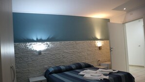 Quarto com parede de pedra, cama com roupa de cama listrada, mesa de cabeceira e luminária fixada na parede.