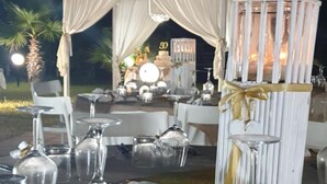 Área de refeições ao ar livre com mesas montadas para um evento formal, com cadeiras brancas, copos e uma estrutura decorativa branca com fita dourada.