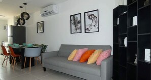 Uma sala de estar moderna com um sofá cinzento, almofadas coloridas, um espaço de refeições com cadeiras e uma mesa, e uma estante preta.