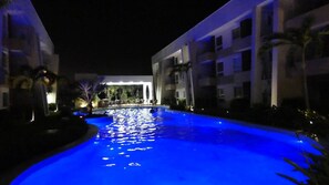Piscina iluminada em azul à noite, cercada por edifícios e palmeiras.