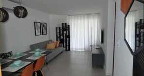 Uma sala de estar moderna com sofá, mesa de jantar e cadeiras. Há uma TV em um móvel, uma estante e um espelho grande.