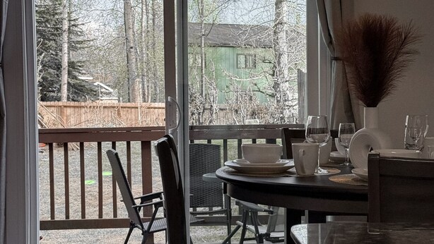 A dining area with a table set for two, a chair, a vase with dried plants, and a view of a green house through a glass door.