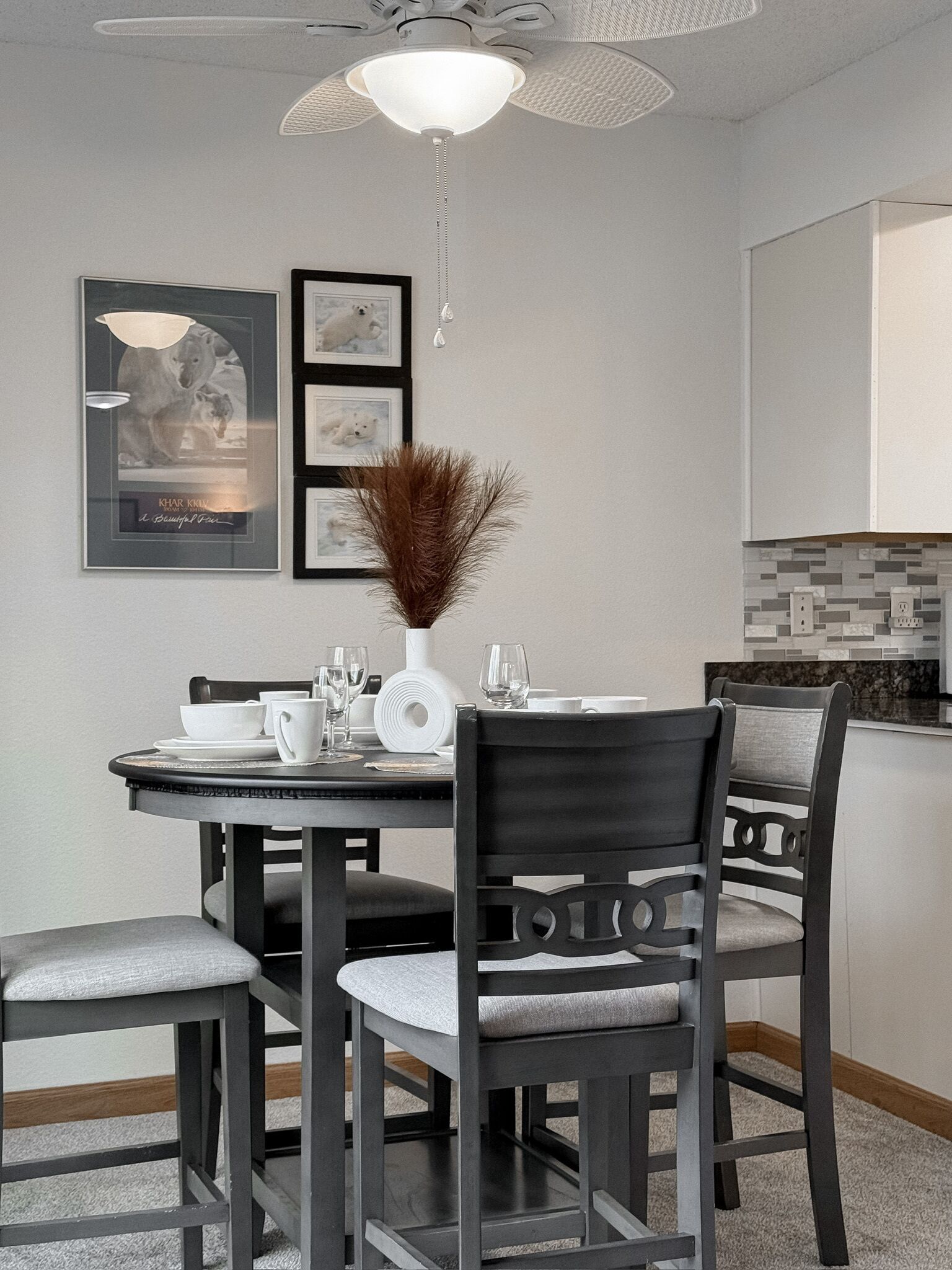 A dining area with a round table, chairs, a ceiling fan, and framed pictures on the wall.