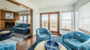 Un salon avec une cheminée, un canapé, deux fauteuils à motifs et une grande fenêtre avec des stores.