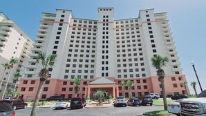 Un hôtel à plusieurs étages, doté d’une entrée principale et entouré de voitures garées et de palmiers.