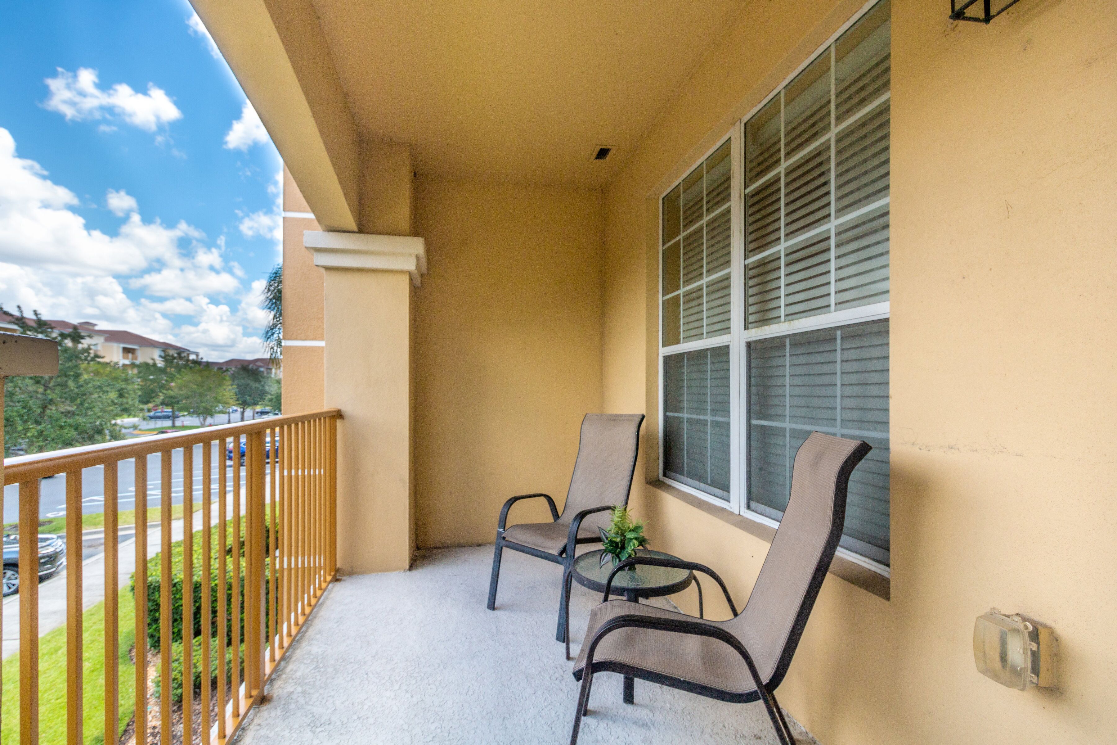 A balcony with two chairs and a small table, a window with blinds, and a view of a street and greenery.