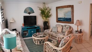 Een woonkamer met een bank, twee fauteuils, een salontafel, een televisie, een ventilator, een plant en een schilderij aan de muur.