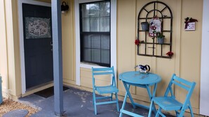 Een veranda met een blauwe deur, een raam met luiken, twee blauwe stoelen, een kleine tafel en een decoratieve metalen rek met potplanten.