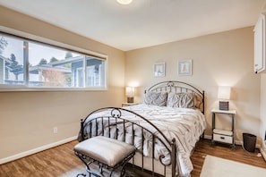 Een slaapkamer met een metalen bed, een raam met zonwering, een nachtkastje met lamp en een klein plankje.