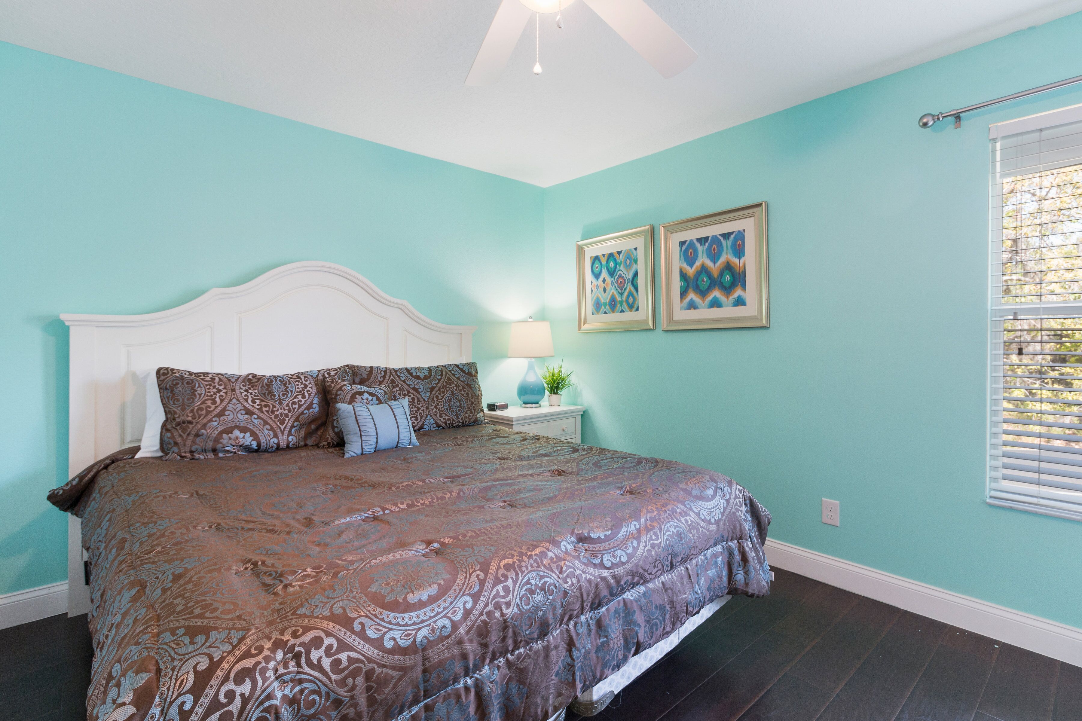 A bedroom with a large bed, a white headboard, two framed artworks on the wall, a bedside table with a lamp, and a window with blinds.
