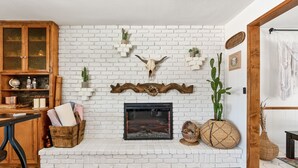 Una chimenea de ladrillo blanco con repisa de madera, rodeada de estantes con plantas y objetos decorativos.
