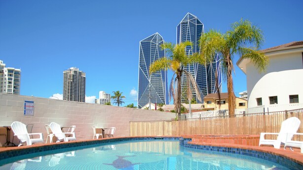 A swimming pool with lounge chairs, a palm tree, and modern buildings in the background. - Gold Coast Inn (Surfers Paradise)