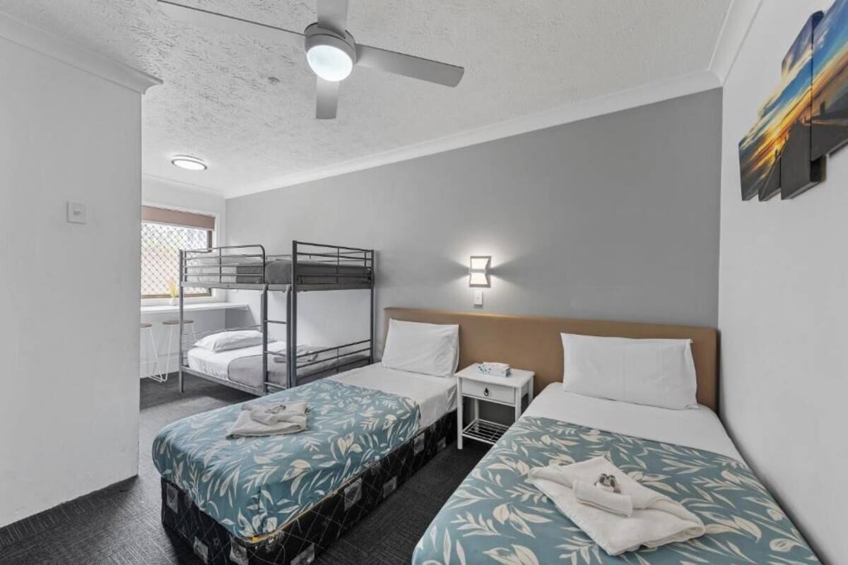 Habitación de hotel con dos camas, una litera, un ventilador de techo, una mesita de noche y una lámpara de pared.
