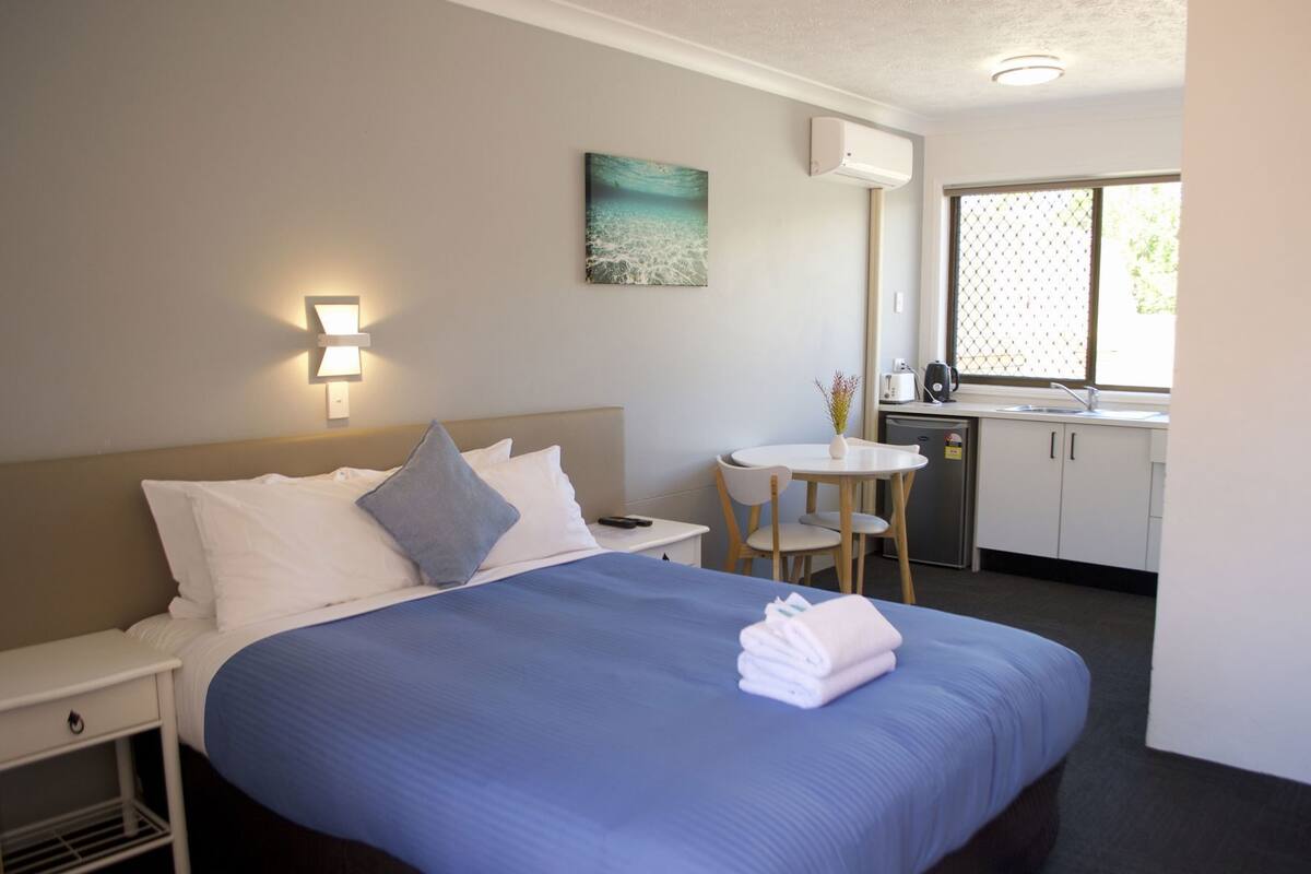 Habitación de hotel con cama, mesita de noche, un pequeño escritorio con silla, ventana y kitchenette.