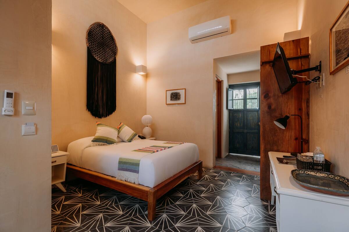 A bedroom with a bed, bedside table, wall-mounted lamp, and a door leading to another room.