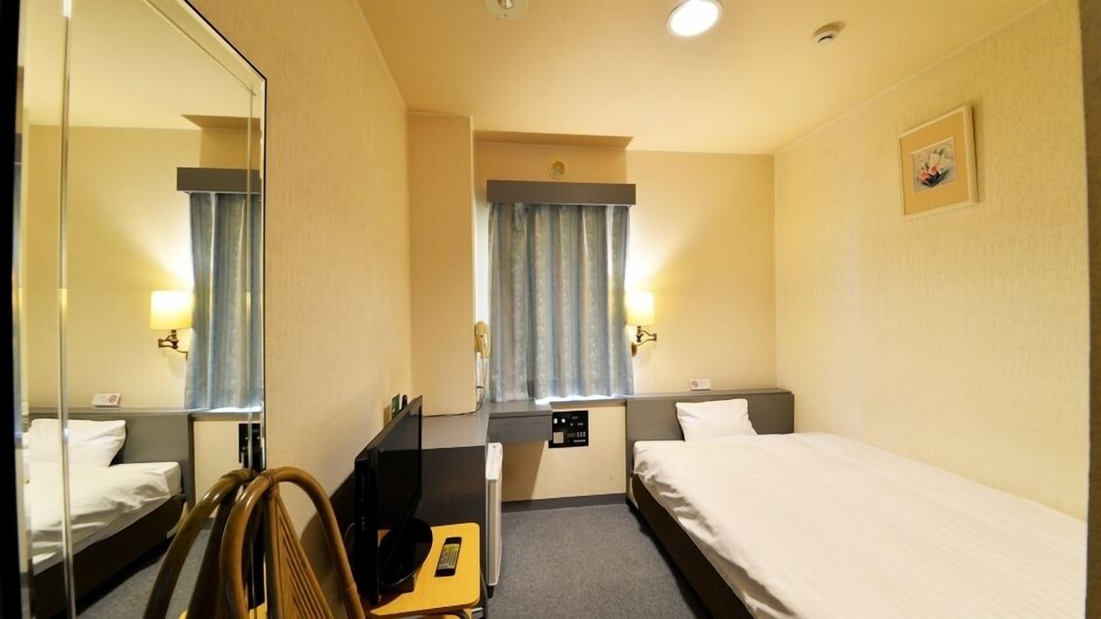 Habitación de hotel con dos camas, un escritorio, una silla, un espejo, una ventana con cortinas y una lámpara de pared.