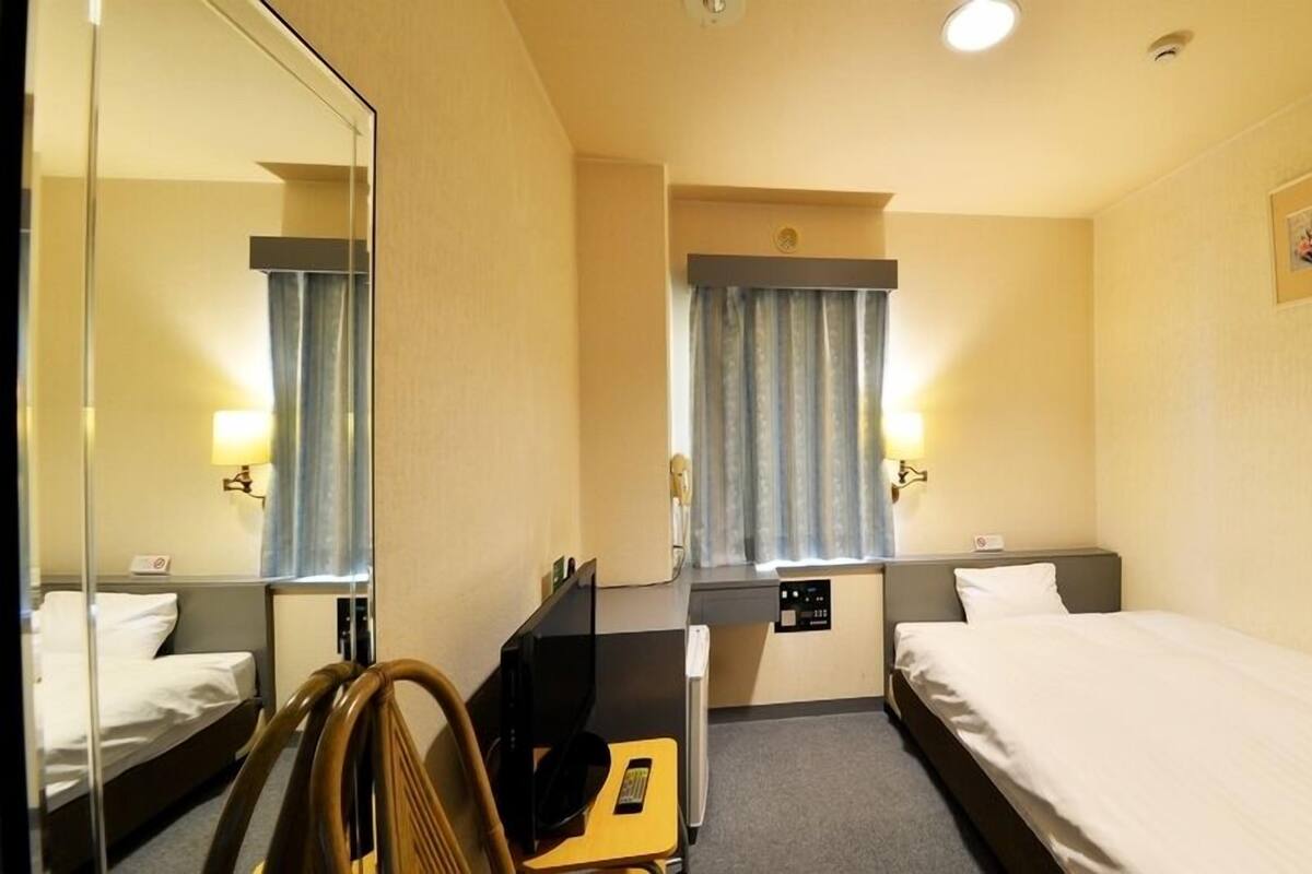 Quarto de hotel com duas camas, uma mesa, uma cadeira, um espelho, uma janela com cortinas e uma luminária na parede.
