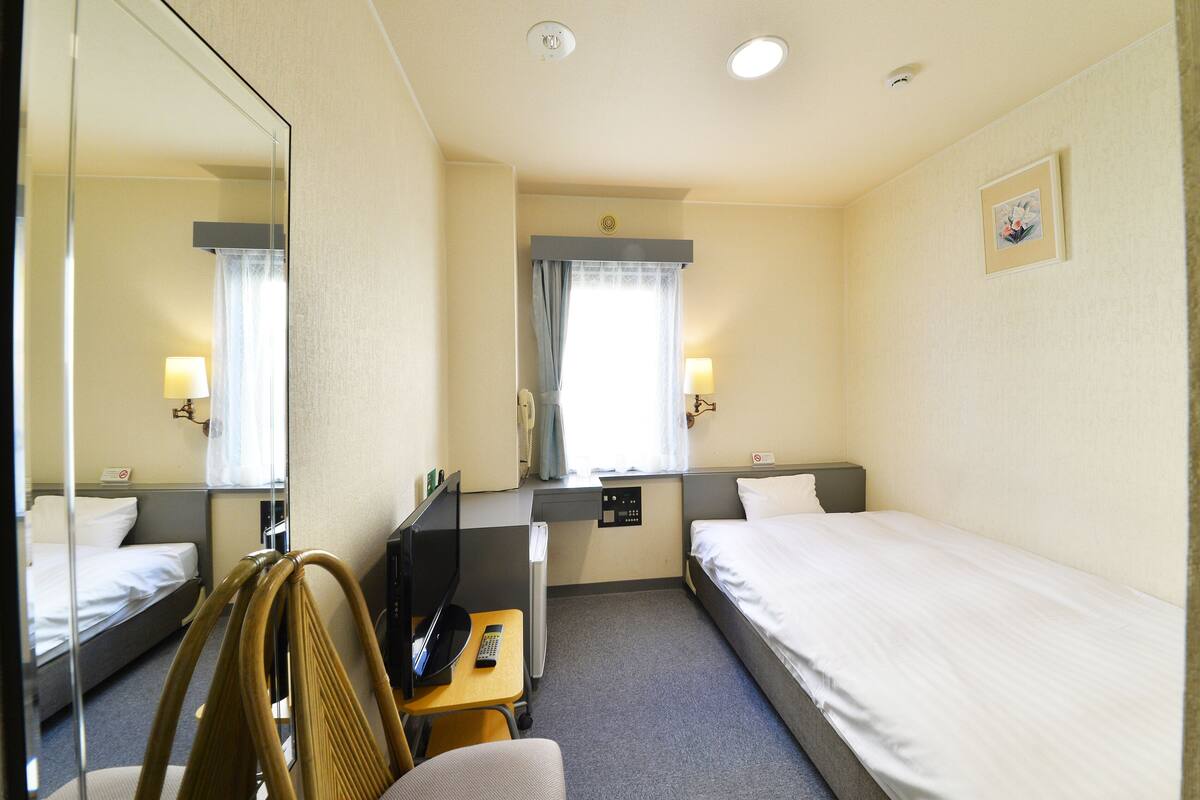 Quarto de hotel com uma cama de solteiro, uma escrivaninha com cadeira, televisão e espelho.