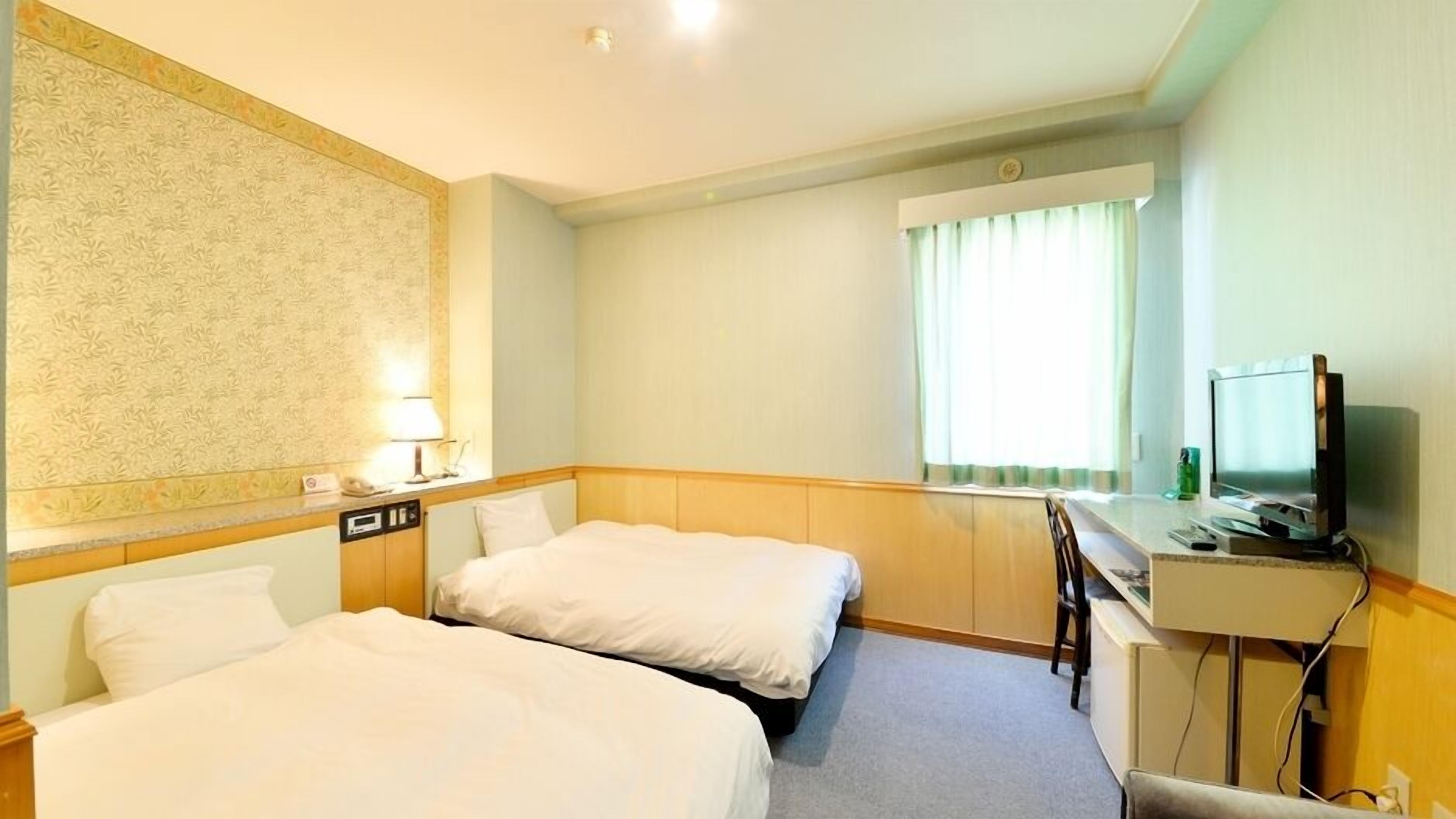 Habitación de hotel con dos camas, un escritorio, televisor y ventana con cortinas.