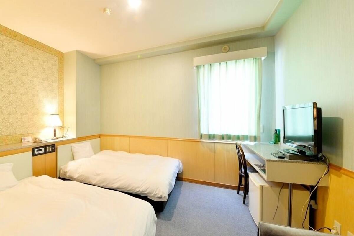 Quarto de hotel com duas camas, uma escrivaninha, televisão e janela com cortinas.