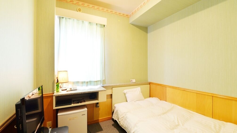 Habitación de hotel con cama, escritorio con computadora, refrigerador pequeño y ventana con cortinas.