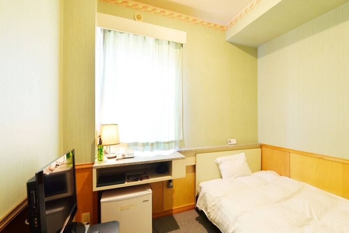 Quarto de hotel com cama, escrivaninha, computador, pequeno frigorífico e janela com cortinas.