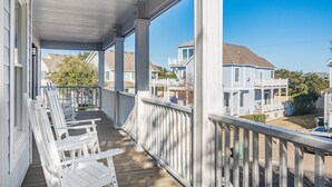 A covered porch with white railings and wooden chairs, overlooking a residential street with houses. - Bluebird: Saltwater Pool, Hot Tub. Golf Community (Corolla)