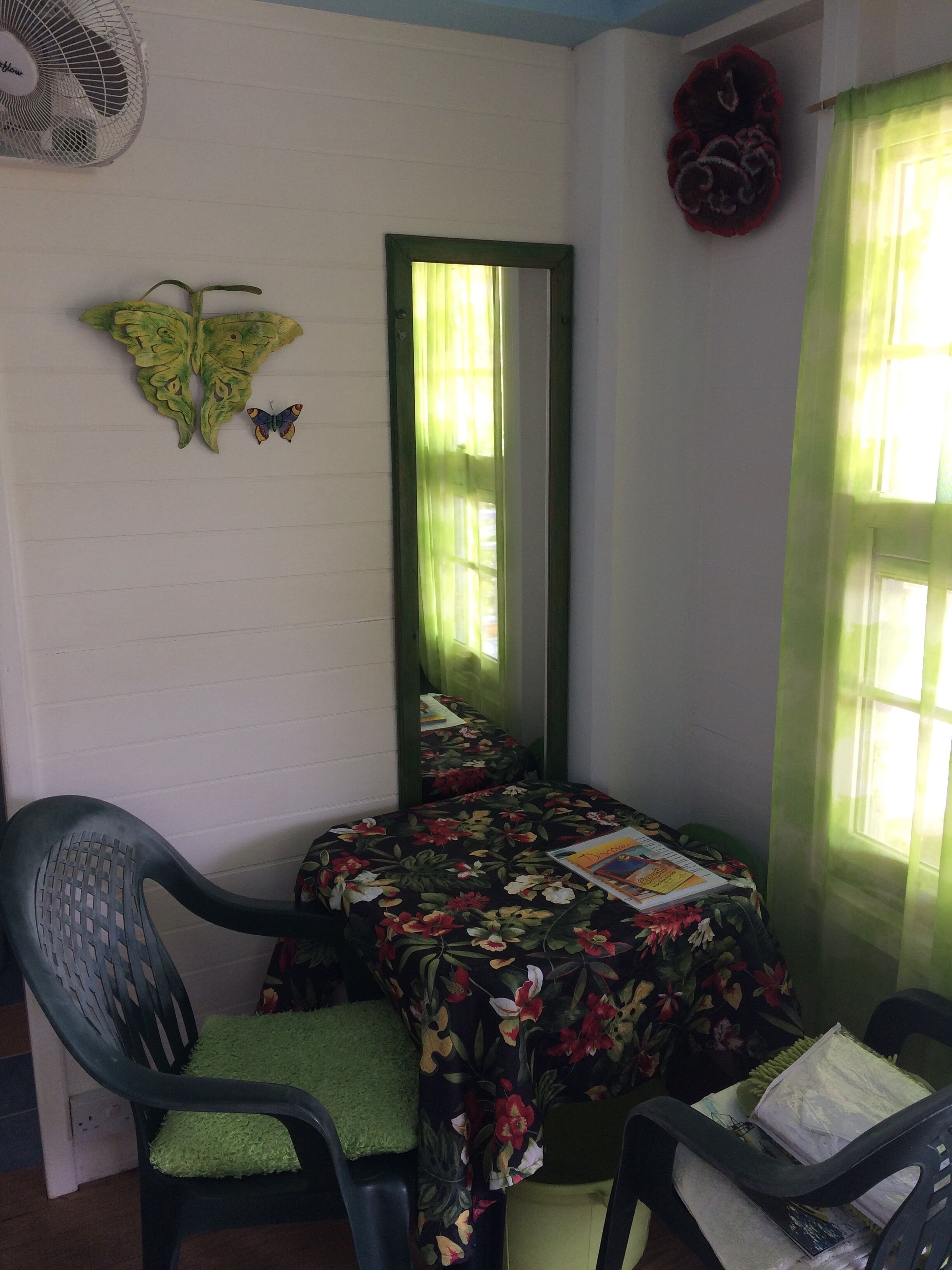 Une pièce d’angle avec un fauteuil vert, une table recouverte d’une nappe à motifs floraux, un miroir et une fenêtre ornée de rideaux verts.