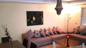 A living room with a sectional sofa, a glass coffee table, a painting on the wall, and a hanging light fixture. - 2 bedroom apartment in agadir (Agadir)