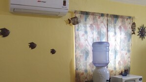 A dining area with a table covered by a patterned tablecloth, chairs, a water dispenser, a microwave, and an air conditioner on the wall.