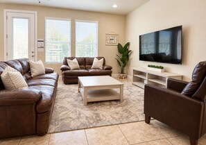 A living room with a brown leather sofa, a matching armchair, a coffee table, and a flat-screen TV mounted on the wall.