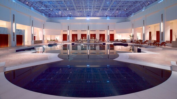 Una amplia piscina interior con techos altos y cubierta de vidrio, rodeada de zonas de estar y un escenario central.