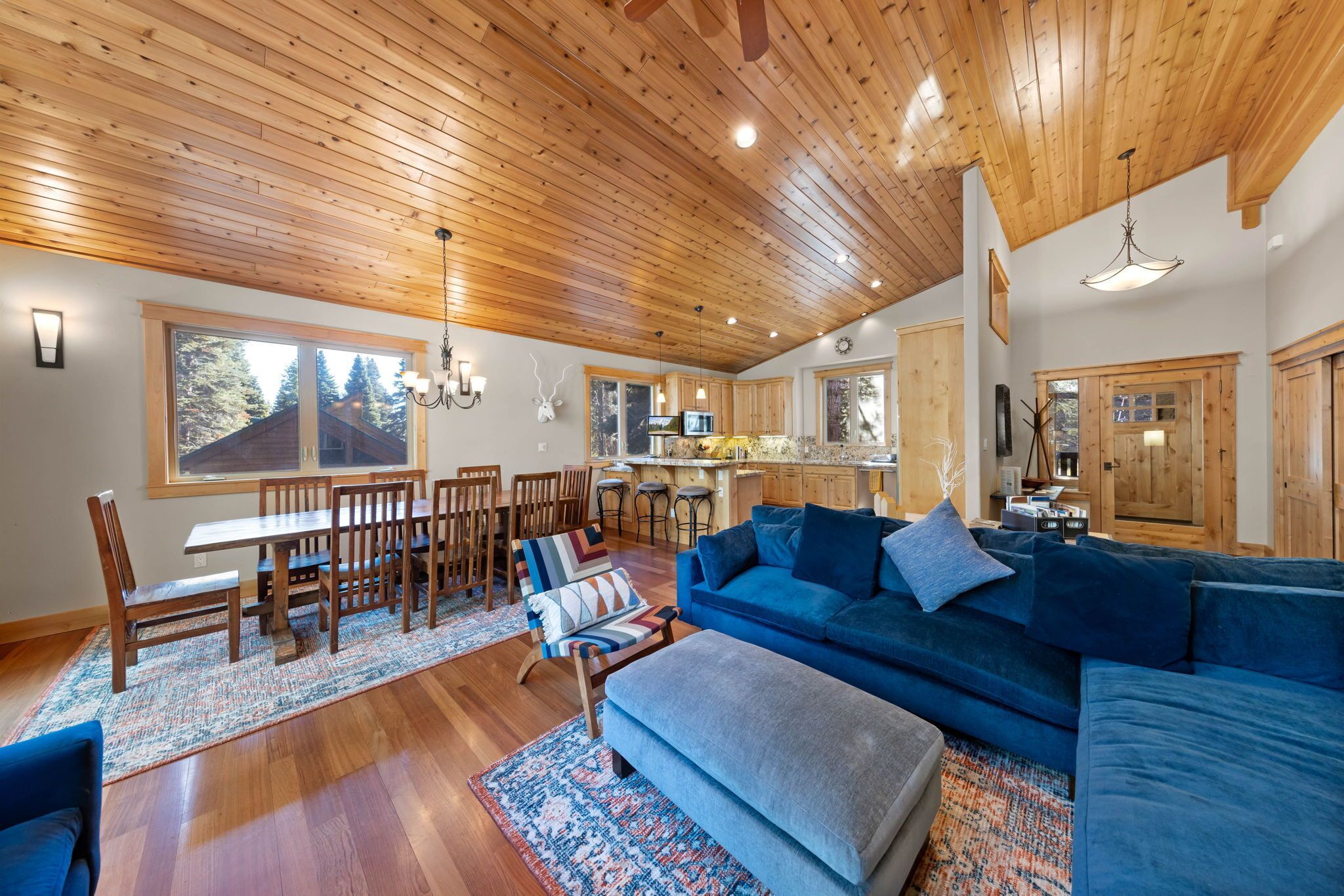A spacious living area with a wooden ceiling, a blue sofa, a dining table with chairs, and a kitchen in the background.
