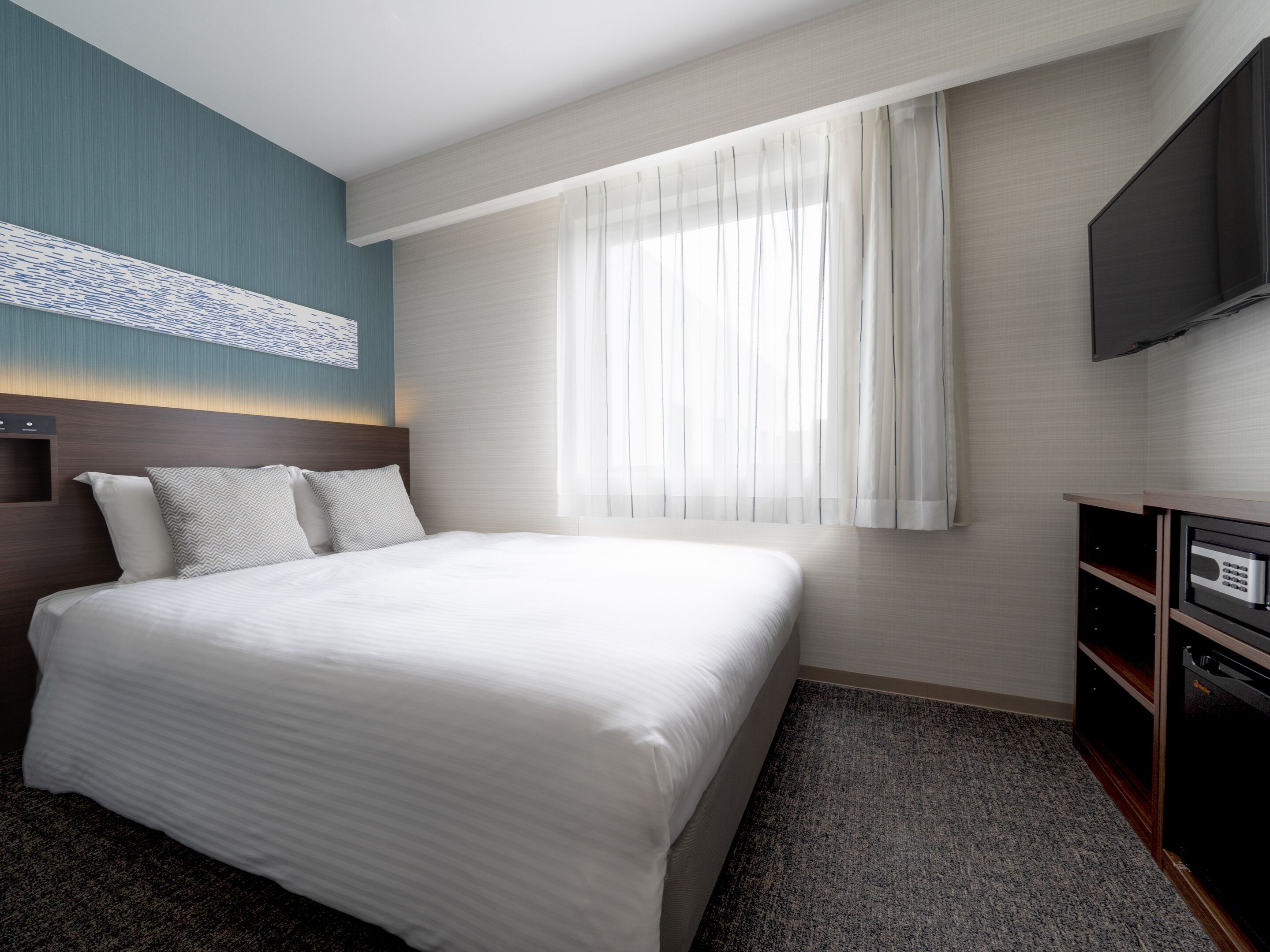 Habitación de hotel con una cama grande, un televisor de pantalla plana montado en la pared, una ventana con cortinas translúcidas y un cabecero de madera.