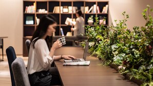 Une personne est assise à une table, un ordinateur portable devant elle, et lit un magazine. Des plantes d’intérieur l’entourent, et une bibliothèque est visible au fond de la pièce.