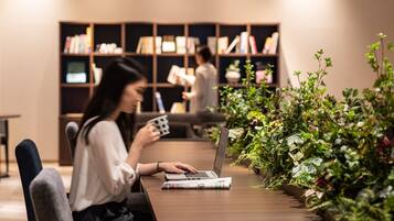 Una persona usando una computadora portátil en una mesa, con una revista, rodeada de plantas de interior, en una habitación con una estantería al fondo.