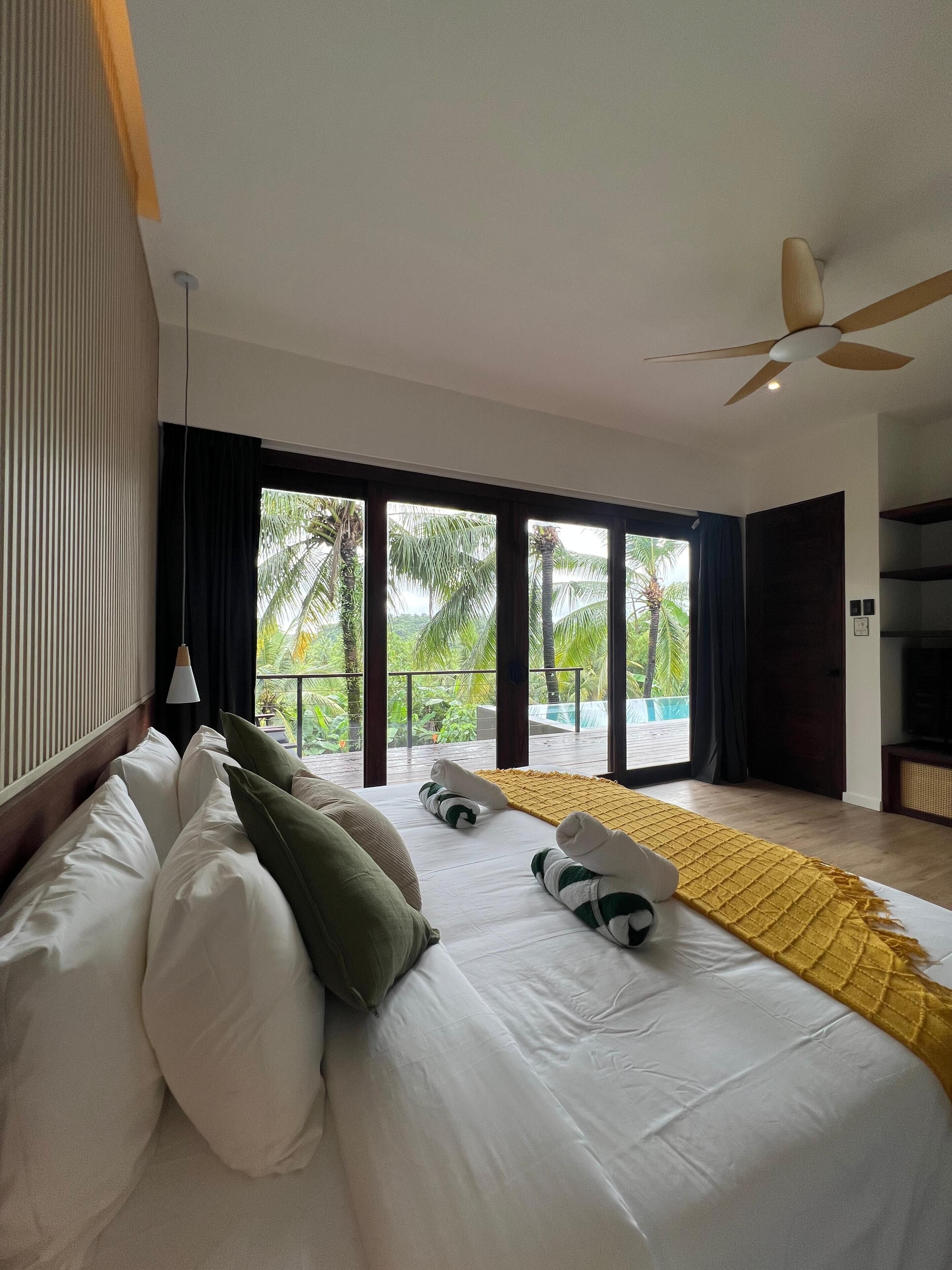 A modern bedroom with a large bed, a ceiling fan, and a view of a pool and palm trees.