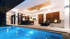 Un área interior de piscina moderna con una piscina cristalina, mobiliario de madera y una zona de comedor.