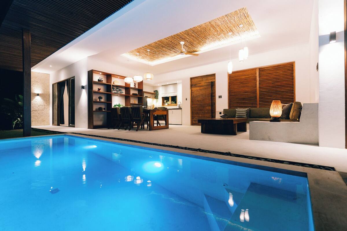 A modern indoor pool area with a clear pool, wooden furniture, and a dining area.
