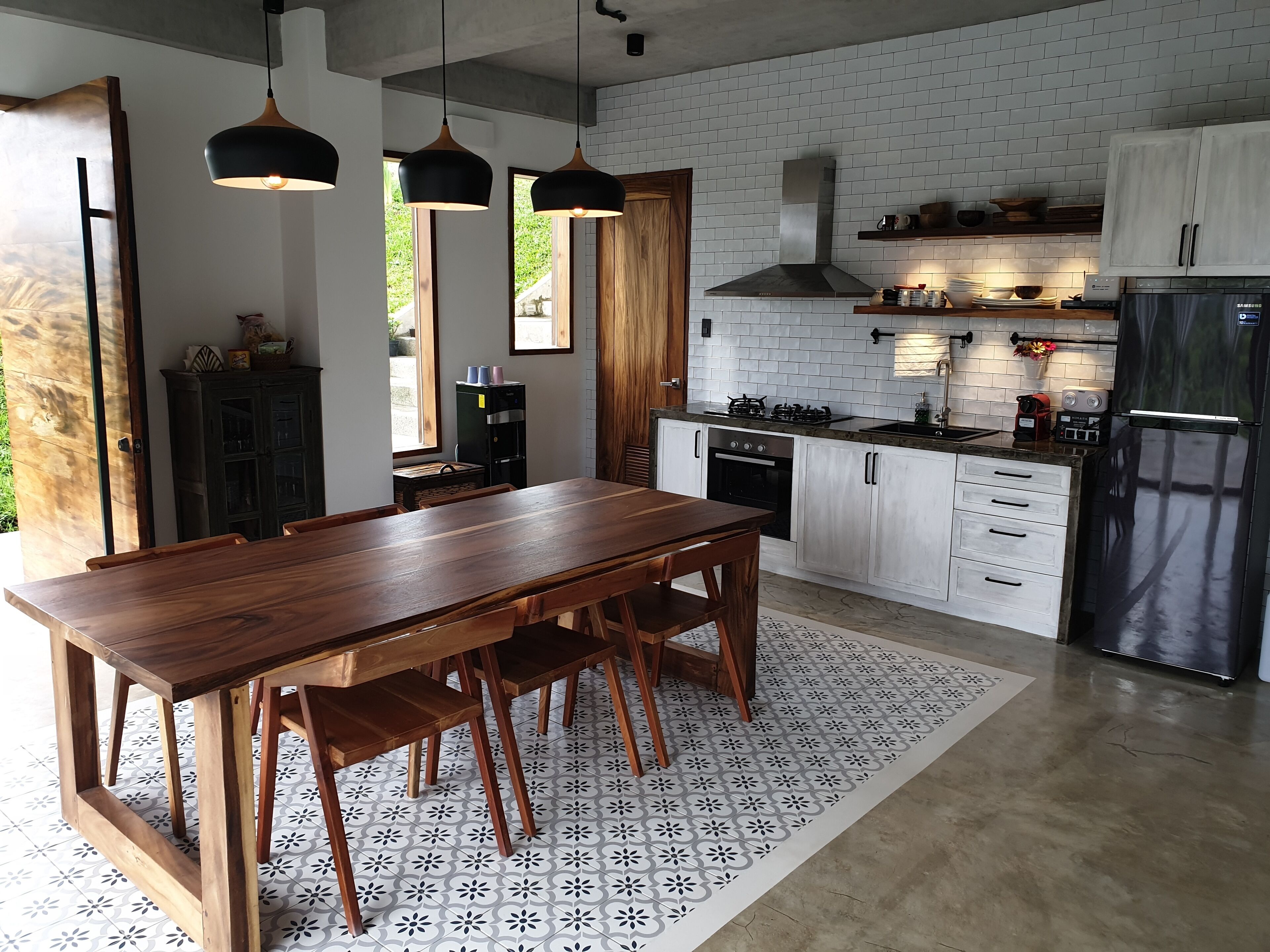 A modern kitchen with white cabinets, a wooden dining table, pendant lights, and a stainless steel refrigerator.