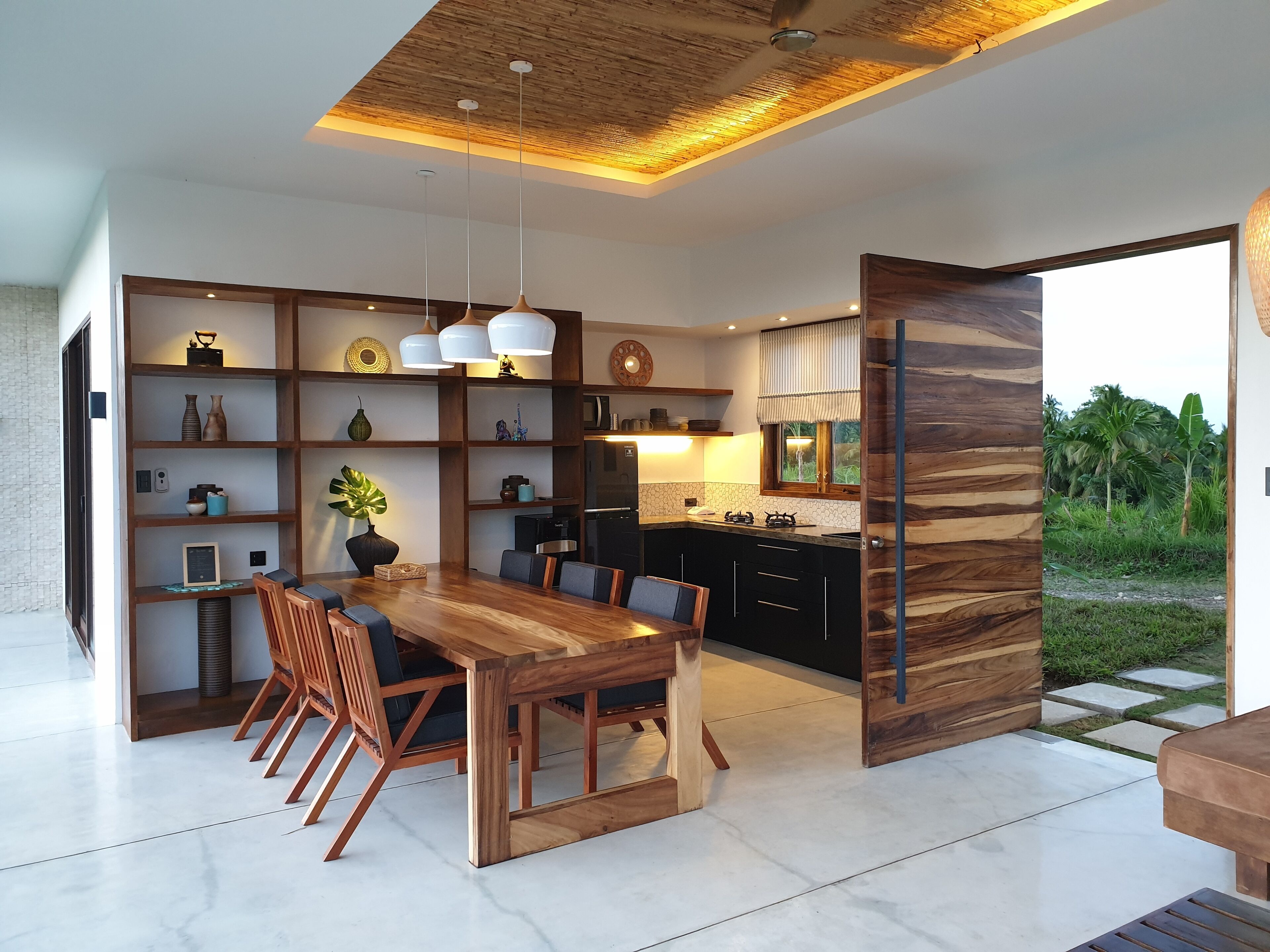 A modern kitchen with a wooden dining table and chairs, built-in shelves, and a view of the outdoors.