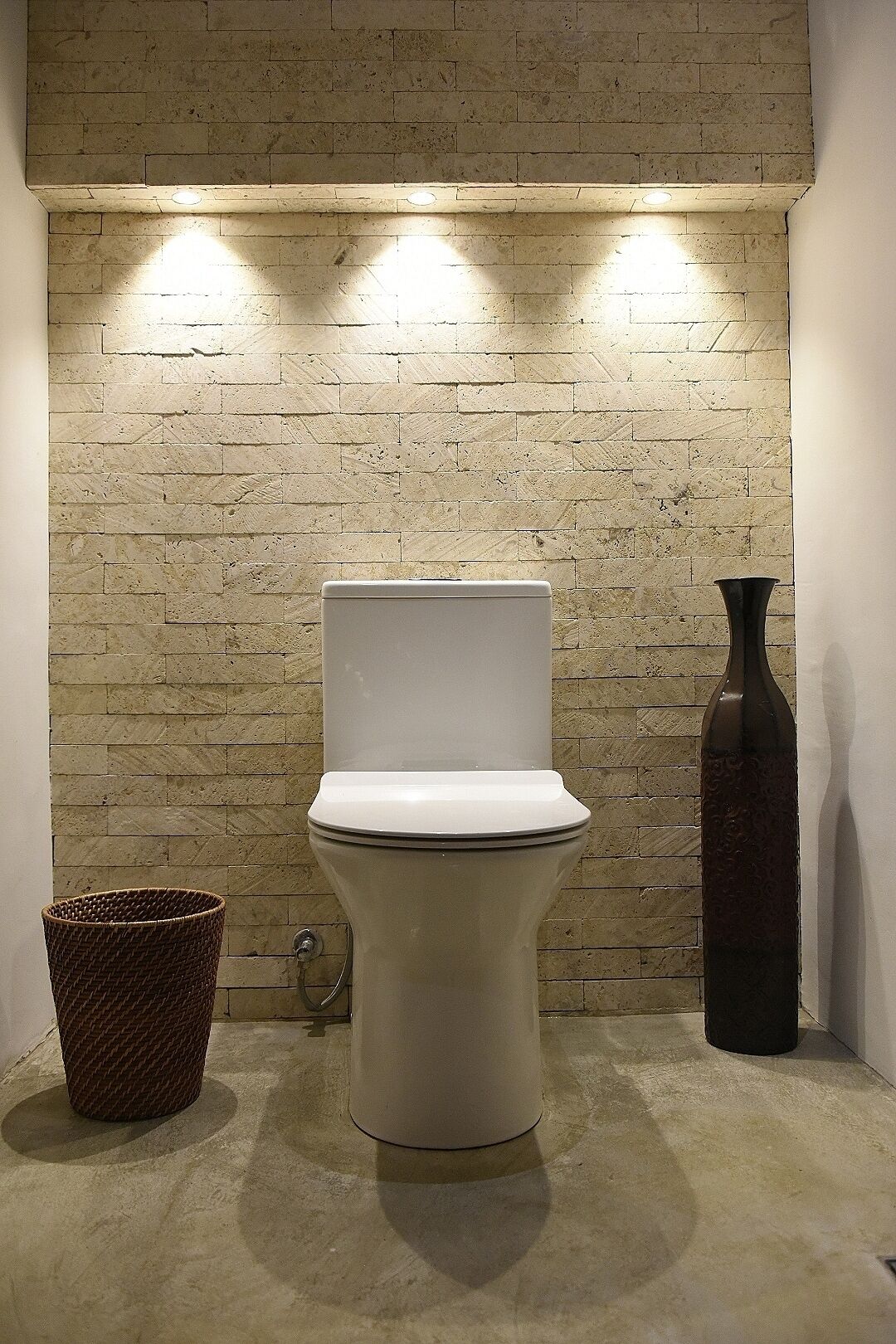 A toilet with a white seat and lid, a dark vase, and a wicker basket against a brick wall with recessed lighting.